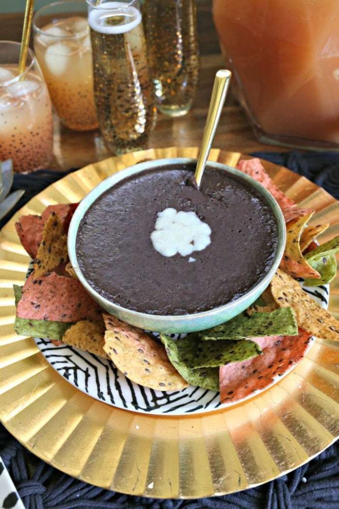 Black bean dip topped with cheese on a gold plate served with tri color tortilla chips.