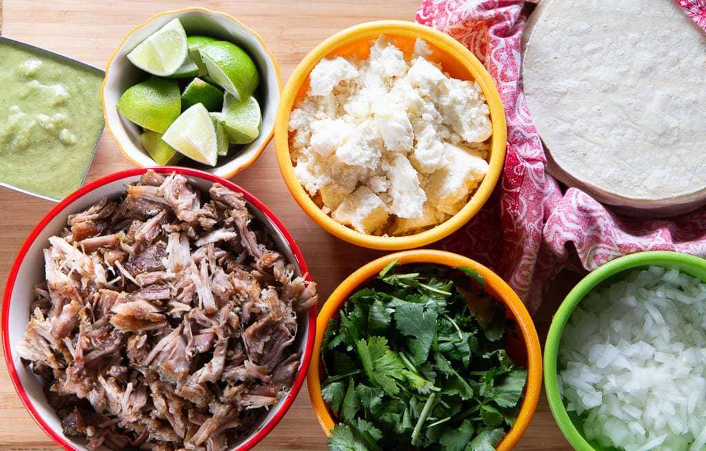 carnitas with garnishes- tortillas, cheese, lime, cilantro and onion